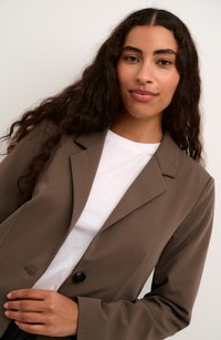 Blazer marrone realizzato in tessuto liscio, con bottone nero, colletto a revers e vestibilità sartoriale, indossato sopra una camicia bianca.