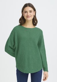 Pull vert ample en maille texturée, doté d'un col bateau et de manches effilées, porté avec un jean.