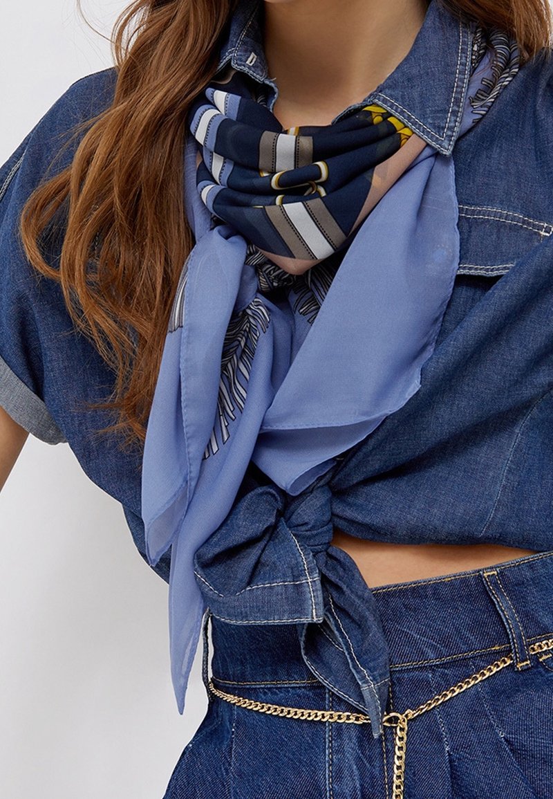 Femme portant des écharpes bleues superposées et une chemise en denim nouée avec un pantalon en denim assorti et une ceinture chaîne dorée.