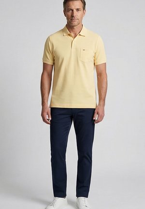 Ein Mann steht, den Blick nach vorn gerichtet, und trägt ein blassgelbes Kurzarm-Poloshirt, marineblaue Hose und weiße Sneaker vor einem schlichten Hintergrund.