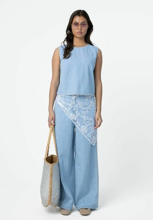 Femme portant un haut brodé bleu clair sans manches et un pantalon large assorti avec un voile en dentelle, tenant un grand sac cabas tissé.