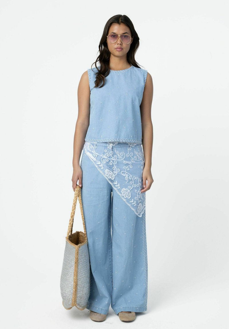 Femme portant un haut brodé bleu clair sans manches et un pantalon large assorti avec un voile en dentelle, tenant un grand sac cabas tissé.