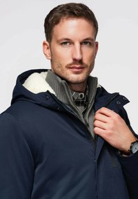 Veste imperméable marine avec une capuche doublée de polaire blanche, portée sur un pull gris à côtes et une chemise à carreaux. Dotée de fermetures éclair et à boutons.