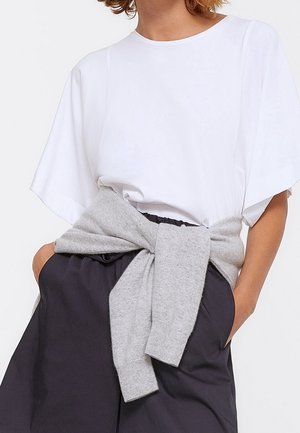 Personne portant une chemise blanche ample avec un pull gris clair noué autour de la taille et un pantalon noir, les mains dans les poches.