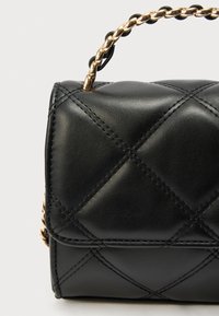 Μαύρη δερμάτινη τσάντα χειρός με καπιτονέ σχέδιο, διαθέτει χρυσή αλυσίδα ως χερούλι και αντίθετη μαύρη ραφή. Συμπαγής ορθογώνιο σχήμα.