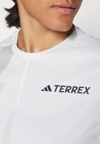 Fehér sportpóló kerek nyakkivágással, fekete "TERREX" logóval a mellkasán, sima, könnyű anyagból készült.
