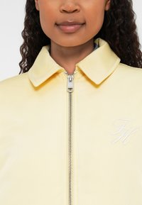 Dzeltena, ar rāvējslēdzēju aizdara jaka ar apkakli, izgatavota no gludas materiāla, ar baltām izšūtām iniciāļiem "FH" kreisajā pusē.