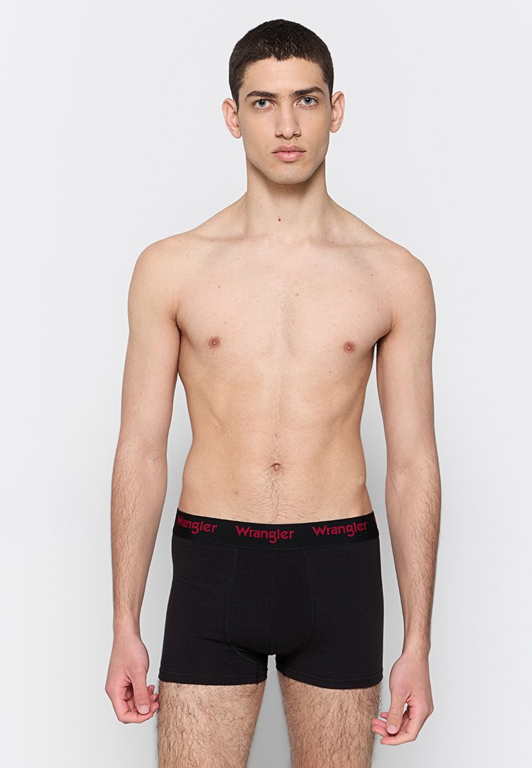 Młody mężczyzna z krótkimi włosami, noszący czarne bokserki z czerwonym logo "Wrangler" na pasie, stojący na jednolitym jasnym tle.