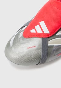 Fotballsko med sølvfarget syntetisk overdel med teksturert finish, rød elastisk stropp og hvite merkevareelementer. "NANOSTRIKE" i rødt.