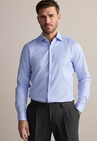 Cămașă albastră deschisă, în carouri, cu guler structurat, mâneci lungi și închidere cu nasturi. Asortată cu pantaloni gri plisati.