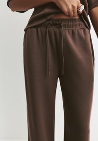 Prim-plan al unei persoane care poartă pantaloni din material textil maro cu șnur și un top asortat, cu o mână ridicându-i puțin cămașa și un inel de argint pe un deget.