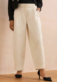 Pantalons larges en ivoire avec des plis doubles, associés à des talons noirs avec des accents en nœud. Texture lisse, design structuré.