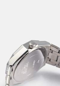 Relógio em aço inoxidável com caixa octogonal, coroa texturizada e pulseira em metal. O fundo da caixa apresenta marcas gravadas.