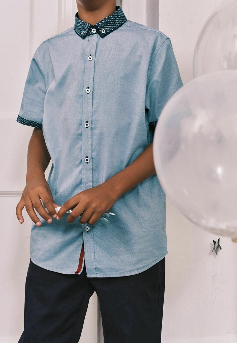 Garçon portant une chemise bleu clair à manches courtes avec une bordure foncée, tenant trois cordons de ballons blancs, avec des ballons transparents en arrière-plan.