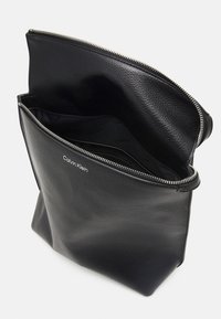 Czarna skórzana torba o teksturowanym wykończeniu, z zamkiem błyskawicznym, otwartą górą i nadrukiem logo wewnątrz. Projekt jest prosty i nowoczesny.
