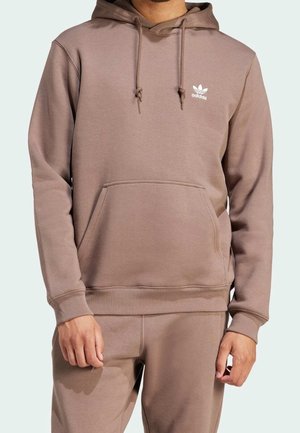 Mand iført lys brun Adidas-hoodie med forlomme og matchende bukser, stående med hænderne afslappet ned langs siden.