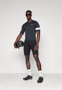 Tenue de cyclisme composée d'un maillot noir à manches courtes avec une accentuation blanche sur les bras, de shorts noirs et de chaussures de cyclisme noires. Tient un casque noir.