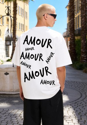 Personne blonde portant des lunettes de soleil noires et une chemise blanche avec "AMOUR" imprimé plusieurs fois en noir, debout dans une rue ensoleillée avec des palmiers.