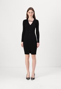 Fekete bordázott kötött ruha hosszú ujjakkal, V-nyakkal, gombos elejével és térd hosszúságú szegéllyel. Egy díszítő pánt részlet található a nyak körül.