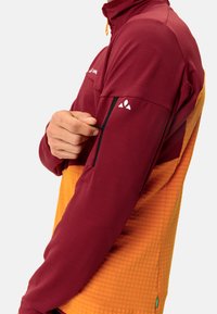 Rote und orange langärmelige Jacke mit einer Reißverschlusstasche am Arm, strukturiertem Stoff und hohem Kragen. Logo auf der Brust und am Ärmel.