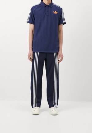 Mann trägt marineblauen Adidas-Trainingsanzug mit weißen Streifen an Ärmeln und Hosen, weiße Sneakers, steht vor einfarbigem Hintergrund.