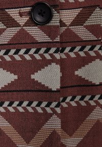 Mörk knapp på tyg med geometriska mönster i brunt, beige, svart och vitt, med trianglar och zickzacklinjer.