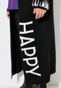 Cardigan en tricot noir avec un grand texte blanc indiquant "HAPPY". Les manches présentent un design rayé noir et blanc et sont côtelées aux poignets.