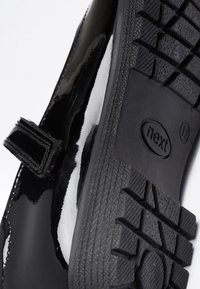 Black patent leather shoe with velcro strap shows textured black rubber sole with "next" logo and size 12 marking.