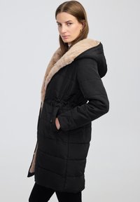 Černý prošívaný kabát s límcem ze světlého umělého kožešiny, stahovací pas a boční kapsy. Obsahuje kapuci a prošívaný design pro teplo.