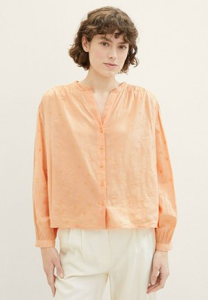 Blouse de couleur pêche avec un col en V, fermeture par bouton et manches longues. Présente un motif texturé et des détails froncés aux épaules.