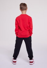 Felpa rossa con maniche lunghe, pantaloni cargo neri e sneakers rosse e bianche. L'outfit presenta un taglio comodo e uno stile casual.