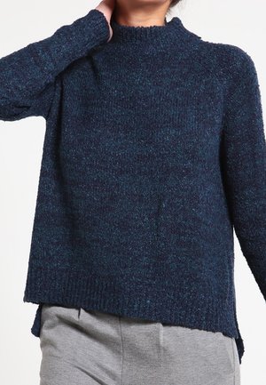 Osoba ubrana w ciemnoniebieski teksturowany sweter z wysokim kołnierzem i jasnoszare spodnie, ręka dotykająca tylnej części szyi.