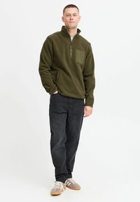 Olive green fleece pullover with a quarter zip, side pockets, and a chest pocket, paired with black jeans and grey sneakers.