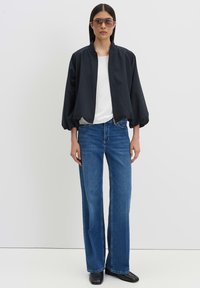 Woman wearing oversized black jacket, white top, blue jeans, black shoes, and sunglasses standing against a plain white background.
