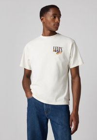 Kreminės spalvos trumpomis rankovėmis medvilnės marškinėliai su daugiaspalviu "LEVI'S" logotipu kairėje krūtinės pusėje, derinami su mėlynais džinsais.