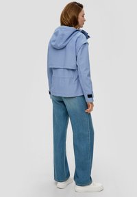 Jasnoniebieska wodoodporna kurtka z kapturem, zapięciem w mankietach i podwójną warstwą, zestawiona z wysokim stanem prostymi niebieskimi dżinsami i białymi trampkami.