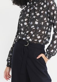 Kwiatowa bluzka w kolorze czarnym z białymi i niebieskimi wzorami, z długimi rękawami i wysokim dekoltem. Połączona z ciemnymi spodniami z wysokim stanem.