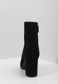 Bottine en suède noir avec un talon haut et large. Présente une fermeture éclair latérale et une texture lisse, avec un design minimaliste et des accents subtils.