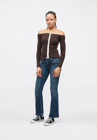 Cardigan côtelé marron à épaules dénudées avec fermeture à boutons blancs, associé à un jean évasé bleu et des baskets basses noires.