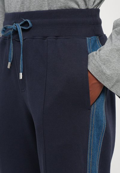 En person iført marineblå joggebukser med denim sidestykker og en knyttet denim snor, hånden i høyre lomme, grå langermet topp synlig.