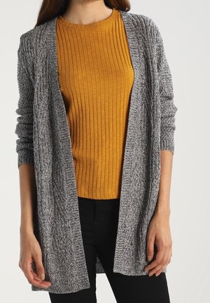 Vrouw draagt een mosterdkleurige geribde trui en een lange, open grijze gebreide cardigan met een zwarte broek tegen een effen achtergrond.