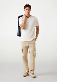 Weißes T-Shirt mit kurzen Ärmeln, beige Cargohosen mit Seitentaschen und lässige Turnschuhe an einem Modell, das eine Jeansjacke hält. Einfaches, modernes Design.