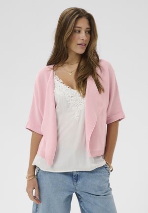 Hosszú barna hajú nő rózsaszín rövid ujjú kardigánt, fehér csipkés szegélyű felsőt, kék farmert, arany karkötőket és rétegezett nyakláncokat visel, és jobbra néz.