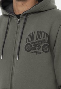 Sudadera con cremallera gris con logo negro que presenta un gráfico de motocicleta. El material parece suave, con una textura lisa y cordones en contraste.