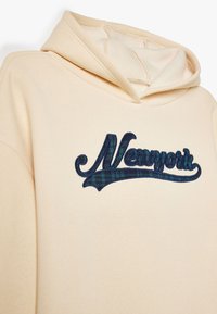 Sudadera cremosa con un logo bordado "New York" texturizado en azul marino y verde. La tela es suave, con un corte relajado y capucha adjunta.