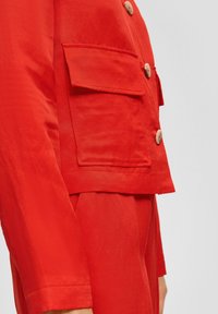 Veste rouge légère avec une coupe structurée, dotée d'une poche poitrine, de boutons en bois et de poignets repliés. Texture lisse.