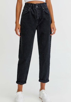 Vrouw draagt zwarte denim jeans met hoge taille en omgeslagen pijpen, gecombineerd met witte sneakers, staand tegen een effen lichte achtergrond.