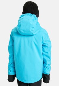 Veste imperméable bleue avec capuche, présentant une texture lisse, des poignets élastiques, et des gants noirs, portée avec un pantalon noir.