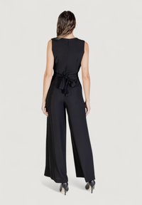 Mono negro con piernas anchas, diseño sin mangas y detalle de cintura anudada. Tejido suave con cierre de cremallera en la parte posterior. Se calzan zapatos de tacón alto.