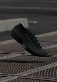 Melnā perforētā Nike Air sporta apava, kas karājas virs metāla režģa virsmas ārējā pilsētas vidē.